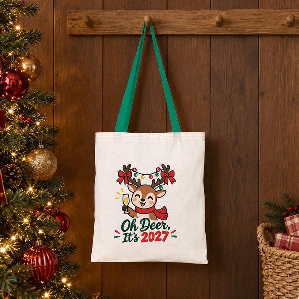 Green handle cotton tote bag with an Oh Deer, It's 2027 reindeer print hanging on a wooden peg rail beside a decorated Christmas tree
