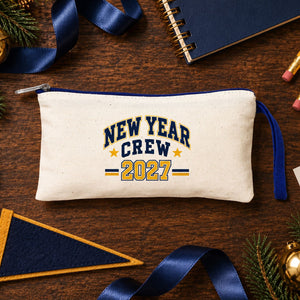 Cream pencil case with a navy blue zipper and a New Year Crew 2027 varsity print on a walnut desk with a notebook, pencils and gold star stickers