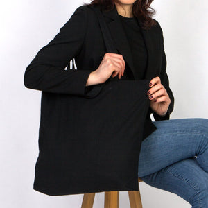 Person wearing a black blazer over a black top and blue jeans, sitting on a wooden stool against a white background.