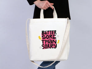 Person holding a tote bag with 'Better Sore Than Sorry' text on a plain background